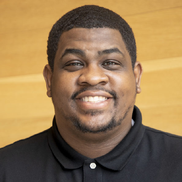 A three-quarter informal portrait of Dennis, he is standing in a gym, smiling towards the camera